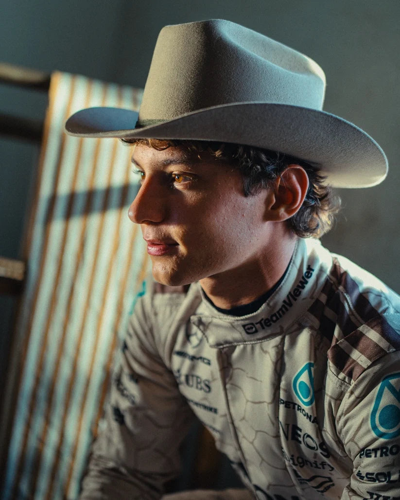 Andrea Kimi Antonelli wearing a cowboy hat and racing suit with sponsor logos, sitting on a wooden bench.