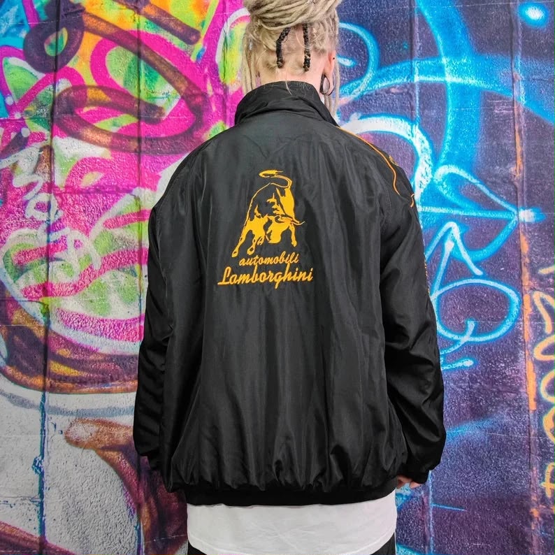 Person wearing a Lamborghini Formula 1 Style Racing Jacket with Lamborghini logo against a colorful graffiti wall.